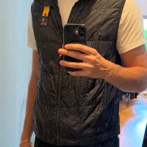 Svart dunväst från Parajumpers XL - Säljer en svart dunväst från Parajumpers i storlek XL. Västen har quiltad design, dragkedja framtill, två sidofickor med dragkedja och ikonisk gul detalj vid kragen. Perfekt för lager-på-lager och snygg streetstil. Passar perfekt om du är runt 180-190! 