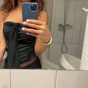 Svart axelbandslös tubtopp med knyt - Snygg svart axelbandslös tubtopp med stor knytning framtill. Gjorde av satin. Sidan är egentligen en drahekedja men som gått sönder och jag behövt sytt ihop som blivit ett litet hål där man kan lägga in bandet om man istället vill ha bandet kortare 