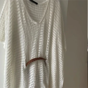 Vit stickad kortklänning med bälte - Superfin vit stickad kortklänning med luftig struktur och v-ringning. Klänningen har ett brunt flätat bälte i midjan som ger en snygg siluett. Perfekt för varma dagar och enkel att styla med sandaler eller sneakers.