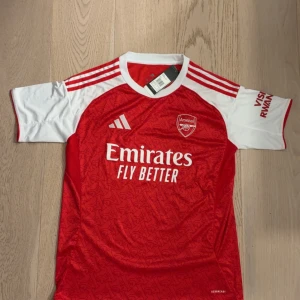 Arsenal hemmatröja 25/26 - Säljer en officiell Arsenal hemmatröja från Adidas i storlek M. Tröjan är röd med vita ärmar och har vita detaljer samt klubbmärke och sponsortryck på bröstet. Tillverkad i lätt och ventilerande material med subtilt mönster i tyget. Perfekt för dig som älskar fotboll och Arsenal.