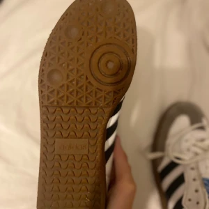 Adidas Samba sneakers og - Adidas Samba sneakers med klassisk design, vita med grå mockadetaljer och svarta ränder på sidorna. Skorna har snörning, platt gummisula och rund tå. Märkets blå logga syns på tungan. Perfekt för dig som gillar retrostil och sportig vibe. Har bara använt som inomhus skor på idrott. Aldrig utomhus.