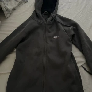 Mörkgrå hoodie från McKenzie - Mörkgrå hoodie från McKenzie med dragkedja framtill och huva med snörning. Tröjan har lång ärm och en enkel, clean design med liten logga på bröstet. Perfekt för chill dagar eller när du vill ha en avslappnad stil.
