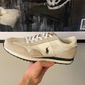 Beige sneakers från Polo Ralph Lauren - Snygga sneakers från Polo Ralph Lauren i beige och vitt med klassisk logga på sidan. Skorna har ovandel i mocka och textil, vita snören och svartvit sula. Perfekta för dig som gillar stilrena och sportiga vibes.