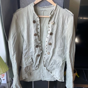 Ljusgrön kavaj med dekorativa knappar - Snygg ljusgrön kavaj med dubbla rader av dekorativa metallknappar och coola axeldetaljer. Jackan har två fickor framtill och råa kanter vid kragen för en avslappnad vibe. Perfekt för dig som vill sticka ut med en unik och trendig look.