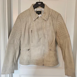 Beige skinnjacka med nitar från Glassly - Snygg beige skinnjacka från Glassly med coola silverfärgade nitar på axlar och ärmar. Jackan har asymmetrisk dragkedja, flera fickor med zip och klassisk krage. Perfekt för dig som vill ha en edgy look med detaljer som sticker ut.