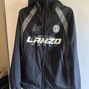 Lorenzo world vindjacka - Svart vindjacka med grå detaljer och tryck från LRNZO World. Jackan har huva, hel dragkedja och sportig design med logga och grafiska element på bröstet. Perfekt för dig som gillar streetwear och vill ha en snygg jacka med attityd.