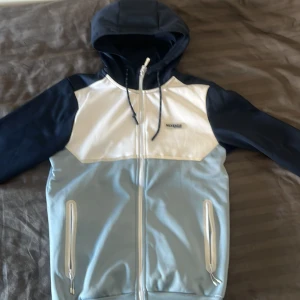 Blå och vit vindjacka från McKenzie - Säljer en snygg vindjacka från McKenzie i blått, vitt och ljusblått. Jackan har huva med dragsko, hel dragkedja framtill och två fickor med dragkedja. Perfekt för dig som gillar sportig stil och vill ha något lätt att slänga på sig.