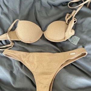 Beige bikini från Aim'n med snörning - Snygg beige bikini från Aim'n med vadderad bygel-bh och knytband i sidorna. Bikinitrosan har en klassisk passform och diskret Aim'n-logga i metall. Materialet är stretchigt och bekvämt, perfekt för strandhäng eller pool.