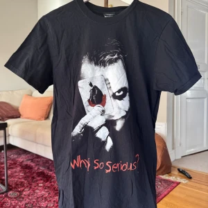 Svart Joker t-shirt Why So Serious - Cool svart t-shirt med tryck av Jokern från Batman och texten 'Why so serious?' i rött. Motivet visar Jokern som håller upp ett spelkort med Batman på. T-shirten har rund hals och korta ärmar, perfekt för dig som gillar film och serier.