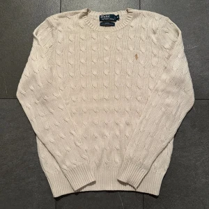 Vit kabelstickad tröja Polo Ralph Lauren - Snygg kabelstickad tröja från Polo Ralph Lauren i storlek M. Färgen är vit och materialet är 100% silke, vilket gör den riktigt mjuk och lyxig. Tröjan har långärmad passform, ribbade muddar och den klassiska broderade loggan på bröstet.