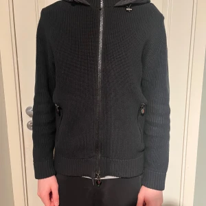 Svart Moncler pufferjacka med huva - Svart pufferjacka från Moncler med stickade ärmar och quiltad rygg. Jackan har huva med snörning, två sidofickor med dragkedja och Moncler-logga på vänster ärm. Snygg, stilren och perfekt för kyliga dagar.