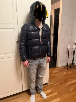 Svart pufferjacka från Save The Duck - Säljer en svart pufferjacka från Save The Duck med huva och dragkedja. Jackan är 100% animal free och har en snygg, glansig finish. Fodret är syntetiskt och jackan har Save The Duck-logga på insidan. Perfekt för kalla dagar och har en modern, loose passform. Jag på bilden är 165 