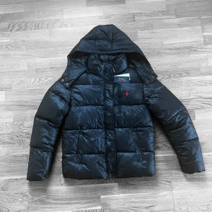 Svart pufferjacka från Polo Ralph Lauren - Svart pufferjacka från Polo Ralph Lauren med huva och dragkedja framtill. Jackan har en röd broderad Polo-logga på bröstet, praktiska fickor och quiltad design. Materialet är glansigt och syntetiskt, perfekt för kalla dagar. Snygg och stilren look.