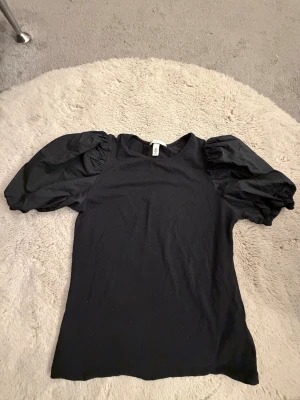 Svart topp med puffärmar från H&M - Säljer en svart topp från H&M med riktigt snygga puffärmar som ger en cool och trendig siluett. Toppen har rund hals och är gjord i mjuk bomull med ärmar i ett lite glansigare tyg. Perfekt för dig som gillar statement-ärmar och stilren look.