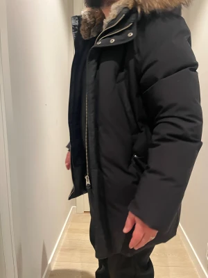 Svart dunjacka med päls på huvan - Säljer en svart dunjacka med fluffig päls på huvan och snygga detaljer som dragkedja och knappar framtill. Jackan är lång och har en loose passform, perfekt för kalla dagar. Huvan är stor och ger extra värme och stil.