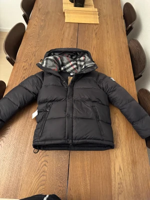Burberry jacka - Svart pufferjacka från Burberry med klassisk logopatch på ärmen och rutigt foder i huvan. Jackan har dragkedja, knappar och två sidofickor med snygga detaljer. Perfekt för kalla dagar och har en riktigt schysst streetstil. Finns äkthetsbevis