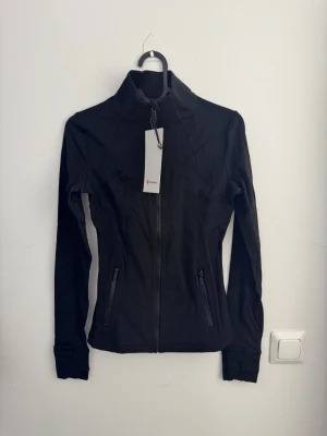 Svart slim fit/tight träningsjacka från Lululemon storlek 4 - Snygg svart träningsjacka från Lululemon med slim fit och dragkedja framtill. Jackan har tumhål vid ärmslut, zip-fickor och ventilerande ryggdetalj. Tillverkad i mjukt, stretchigt Nulu-material med Lycra för extra komfort. Perfekt för aktiva dagar.