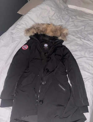 Svart Canada Goose parkas med päls - Svart parkas från Canada Goose med äkta päls på huvan, klassisk logga på ärmen och stora fickor framtill. Jackan har både dragkedja och knappar, samt en varm dunfyllning som gör den perfekt för kalla vinterdagar. Snygg och tidlös modell med lång passform.