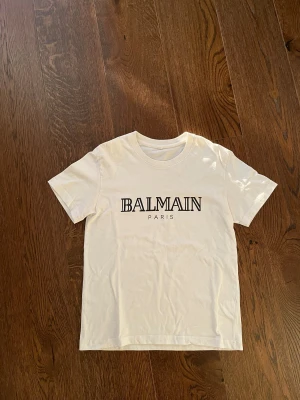 Vit Balmain Paris t-shirt - Snygg vit t-shirt från Balmain Paris med klassisk svart logga framtill. T-shirten har rund halsringning och korta ärmar. Perfekt för dig som gillar stilrena och exklusiva plagg. Gjord i mjuk bomull för skön känsla hela dagen.