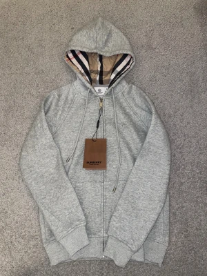 Grå hoodie från Burberry med dragkedja - Snygg grå hoodie från Burberry med klassiskt rutigt foder i huvan och broderad logga på bröstet. Tröjan har snörning i huvan, ribbade muddar och dragkedja framtill. Perfekt för dig som gillar exklusiva streetwear-vibbar. Passar storlek S/XS väldigt liten är den.