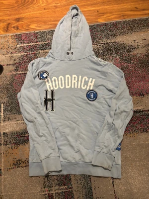 Ljusblå hoodie från Hoodrich  - Snygg ljusblå hoodie från Hoodrich med stor huva, vita tryck och broderade patches på bröstet. Tröjan har en klassisk känguruficka och ribbade muddar vid ärmslut och nederkant. Perfekt för dig som gillar streetwear och vill sticka ut.