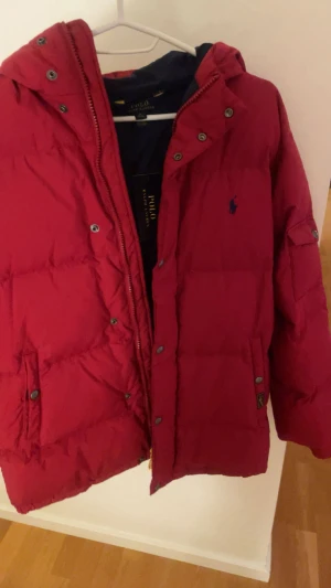 Röd dunjacka Polo Ralph Lauren XL - Snygg röd dunjacka från Polo Ralph Lauren i storlek XL junior. Jackan har quiltad design, tryckknappar och dragkedja framtill samt huva. Klassisk Polo-logga broderad på bröstet. Perfekt för kalla dagar och har en loose passform.