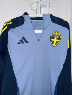Svenska landslaget långärmad fotbollströja - Säljer en långärmad fotbollströja från Adidas med Sveriges landslagsemblem på bröstet. Tröjan är ljusblå med mörkblå ärmar och gula detaljer på axlarna. Materialet är funktionellt och lätt, perfekt för träning eller match. Halv dragkedja framtill och sportig design.