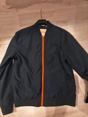 Svart bomberjacka från Jack & Jones - Snygg svart bomberjacka från Jack & Jones med orange dragkedja och ribbade muddar. Jackan har ett coolt meshfoder med tropiskt mönster och två fickor framtill. Perfekt för dig som vill ha en stilren jacka med lite edge.