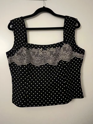 Svart prickig blus med spetsdetalj - Supersöt svart blus från H&M med vita prickar och breda axelband. Framsidan har en bred spetsdetalj i vitt med blommigt mönster som ger en extra touch. Blusen har fyrkantig urringning och är lätt figursydd för en snygg siluett.