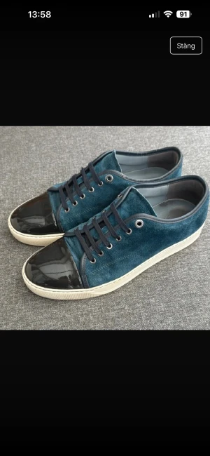 Lanvin blå mocka sneakers - Snygga sneakers från Lanvin i mörkblå mocka med svarta lackade tår och vita gummisulor. Skorna har svarta snören och metallöglor som ger en cool kontrast. Perfekt för dig som gillar unika detaljer och lyxig känsla.