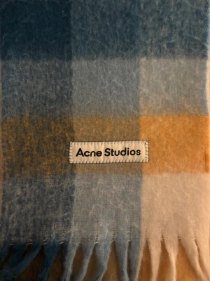Rutig blå och gul halsduk Acne Studios - Snygg och fluffig halsduk från Acne Studios med rutigt mönster i blått, gult och vitt. Materialet är mjuk ull och halsduken har långa fransar i ändarna. Perfekt för att ge din outfit en cool touch och hålla dig varm under kyliga dagar.