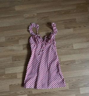 Rutig rödvit kortklänning med volang - Superfin rödvit rutig klänning med volangdetaljer på axelbanden och söt knytning framtill. Klänningen är kort och har en figurnära passform med ärmlös design. Perfekt för sommarens alla tillfällen och har en lätt och luftig känsla.