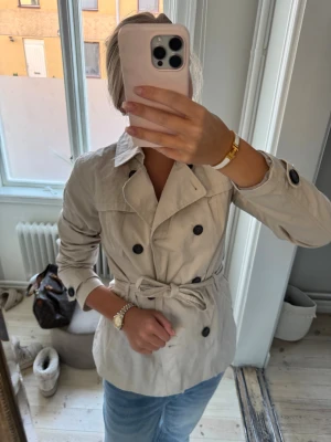 Beige trenchcoat med bälte - Snygg beige trenchcoat med dubbelknäppning och knytskärp i midjan. Jackan har klassisk krage, svarta knappar och långa ärmar med knappdetaljer vid handlederna. Perfekt för dig som gillar stilrena och tidlösa plagg.