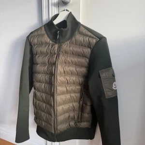 Moncler Cardigan - Snygg olivgrön pufferjacka från Moncler med quiltad front och ärmficka med logotyp. Jackan har dragkedja, ribbad krage och ärmar i stickad ullmix. Perfekt för dig som vill ha en clean och exklusiv look med street-vibe.