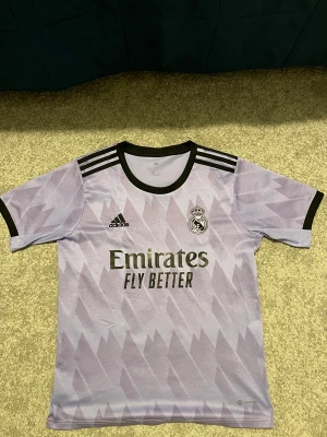 Real Madrid Benzema lila tröja  - Säljer en lila Real Madrid fotbollströja från Adidas med svart tryck, nummer 9 och Benzema på ryggen. Tröjan har korta ärmar, svart krage och svarta detaljer på axlarna. Materialet är lätt och andas, perfekt för träning eller match. Snyggt geometriskt mönster över hela tröjan.