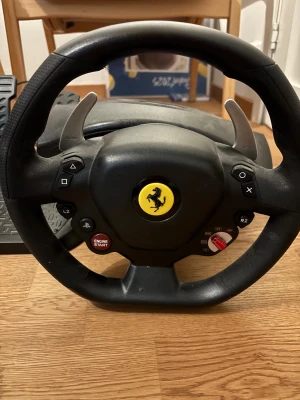 Thrustmaster Ferrari Racing Wheel - Thrustmaster Ferrari Racing Wheel med pedaler, USB-anslutning. Realistisk ratt med Ferrari-logo, växelpaddlar och PlayStation-knappar. Komplett set i gott skick, endast lätt ytslitage. Perfekt för racing på PC och konsol. Använd men fortsatt i gott skick och fungerar galant!