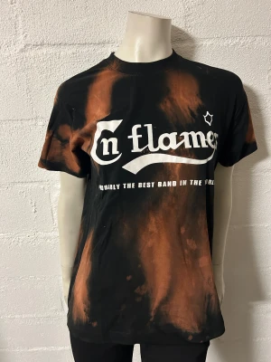 In flames T-shirt med meleringar stl S - Cool svart t-shirt med orangea blekta detaljer och stort In Flames-tryck på bröstet. Rund hals och normal passform. Perfekt för dig som gillar bandmerch och vill sticka ut med en unik look. Storlek S. Helt ny.