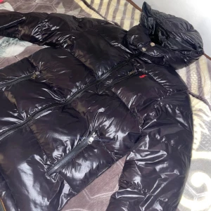 Svart pufferjacka från Polo Ralph Lauren - Snygg svart pufferjacka från Polo Ralph Lauren med glansig finish, huva och flera dragkedjefickor. Jackan har klassisk röd logga på bröstet och är perfekt för kalla dagar. Fodrad insida och hög krage för extra värme.