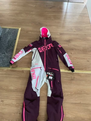 FXR Monosuit Plum/Lt Grey/Elec Pink - Säljer en FXR Ranger Instinct monosuit i färgerna plommonlila, ljusgrå och elektrisk rosa. Dräkten har flera dragkedjor, huva och stora FXR-loggor på bröst, ben och armar. Materialet är slitstark syntet, perfekt för vinteräventyr. Snygg och sportig look med cool färgblock-design.