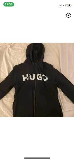 Svart hoodie från Hugo med tryck - Svart hoodie från Hugo med stort vitt tryck på bröstet. Tröjan har huva med snörning, dragkedja framtill och långa ärmar. Materialet är mjukt och känns som bomull, perfekt för chill dagar. Snygg och enkel design med coolt mönstrade bokstäver.