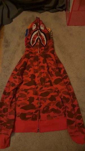 Röd camo hoodie från BAPE Shark - Säljer en ikonisk BAPE Shark hoodie i röd camo med full zip och huva med hajansikte, broderade detaljer och patchar. Hoodien har ribbade muddar, kängurufickor och är tillverkad i mjuk bomull. Perfekt för dig som vill sticka ut med streetwear.