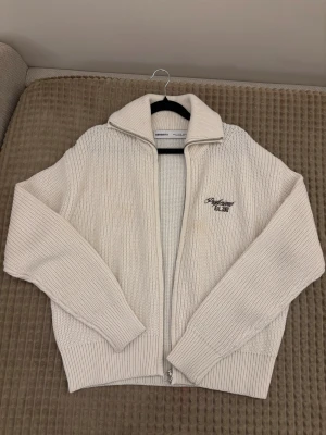 Vit stickad kofta från Bershka - Säljer en vit stickad kofta från Bershka med broderad text på bröstet och dragkedja framtill. Koftan har en hög krage och långa ärmar, perfekt för att hålla värmen. Ribbstickade muddar vid ärmslut och nederkant ger en snygg och chill look.