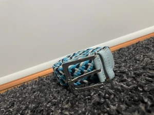 Blått och vitt randigt flätat bälte - Säljer ett snyggt flätat bälte med randigt mönster i blått, vitt och svart. Spännet är i silverfärgad metall och ändarna är i ljusblått syntetmaterial. Perfekt för att ge outfiten lite extra färg och stil.