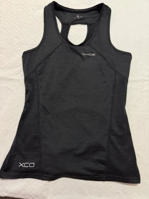 Svart träningslinne från Oxide - Sportigt svart linne från Oxide med XCO-tryck framtill. Modellen är ärmlös med racerback och har en snygg detalj med rosa dragkedja baktill. Materialet är lätt polyester som andas och har en reflekterande rand på ryggen. Perfekt för gymmet eller löprundan.