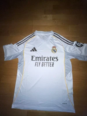 Real Madrid vit matchtröja Adidas S - Säljer en officiell Real Madrid matchtröja från Adidas i slim fit, storlek S. Tröjan är vit med svarta Adidas-ränder på axlarna, gula detaljer och Real Madrid-logga på bröstet. Materialet är lätt och andas, perfekt för träning eller match. Aeroready-teknologi för extra komfort.