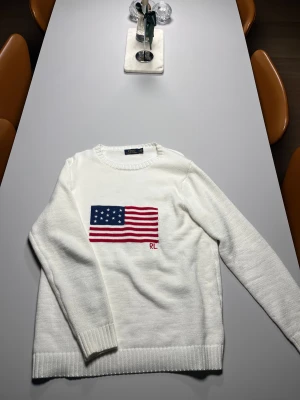 Vit stickad tröja Polo Ralph Lauren - Vit stickad tröja från Polo Ralph Lauren med amerikansk flagga i blått och rött på bröstet samt RL-broderi. Tröjan har rund hals, ribbade muddar och lång ärm. Perfekt för dig som gillar klassisk och clean stil med en twist.