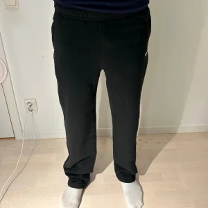 Svarta mjukisbyxor från Nike NOCTA - Svarta mjukisbyxor från Nike NOCTA med vit logga på sidan och broderade detaljer bak. Byxorna har normal passform, elastisk midja och raka ben. Tillverkade i mjukt bomullsmaterial, perfekta för chill eller träning. Snygga fickor på sidorna och stilren design.  Aldrig använda eftersom de är köpta i fel storlek för mig. 