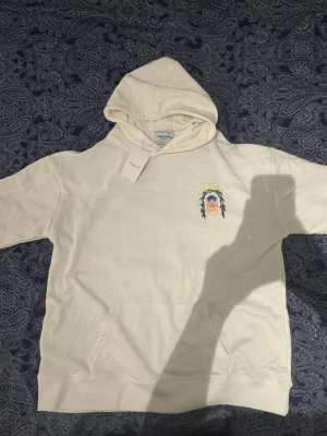 Vit hoodie från Casablanca med tryck - Säljer en vit hoodie från Casablanca med färgglatt tryck av en byggnad och blad på ryggen samt mindre tryck på bröstet. Hoodien har huva, känguruficka och långa ärmar. Perfekt för dig som gillar snygga detaljer och streetwear vibes.