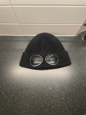 Svart C.P. Company goggle mössa - Svart ribbstickad mössa från C.P. Company med ikoniska goggle-detaljer på uppviket. Mössan är tillverkad i mjukt stickat material och har en klassisk rund form med bred uppvikt kant. Perfekt för dig som vill ha streetstyle-vibe och unik design.