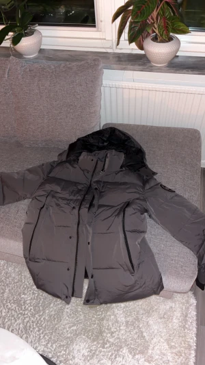 Mörkgrå Canada Goose dunjacka - Säljer en mörkgrå dunjacka från Canada Goose med huva och svarta detaljer. Jackan har lång ärm, dragkedja och knappar framtill samt logotyp-patch på ärmen. Insidan är fodrad i svart och jackan har flera praktiska fickor. Perfekt för kalla vinterdagar.